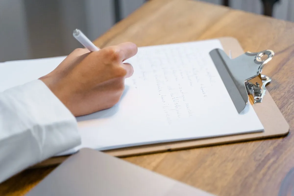 Une main qui écrit au stylo sur un porte-bloc, sur une table en bois.