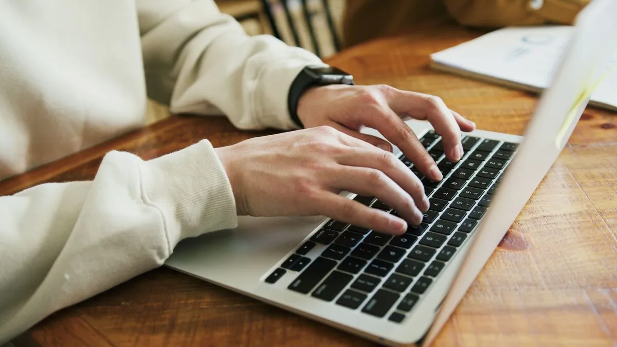 Des mains sur le clavier d’un ordinateur portable posé sur une table en bois.